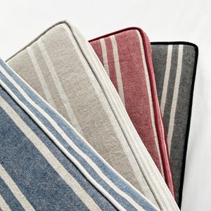 May include: Close-up of four striped cushions in shades of blue, beige, red, and gray. Each cushion has a white border and a different stripe pattern. The cushions are arranged diagonally, showcasing their varying colors and textures.