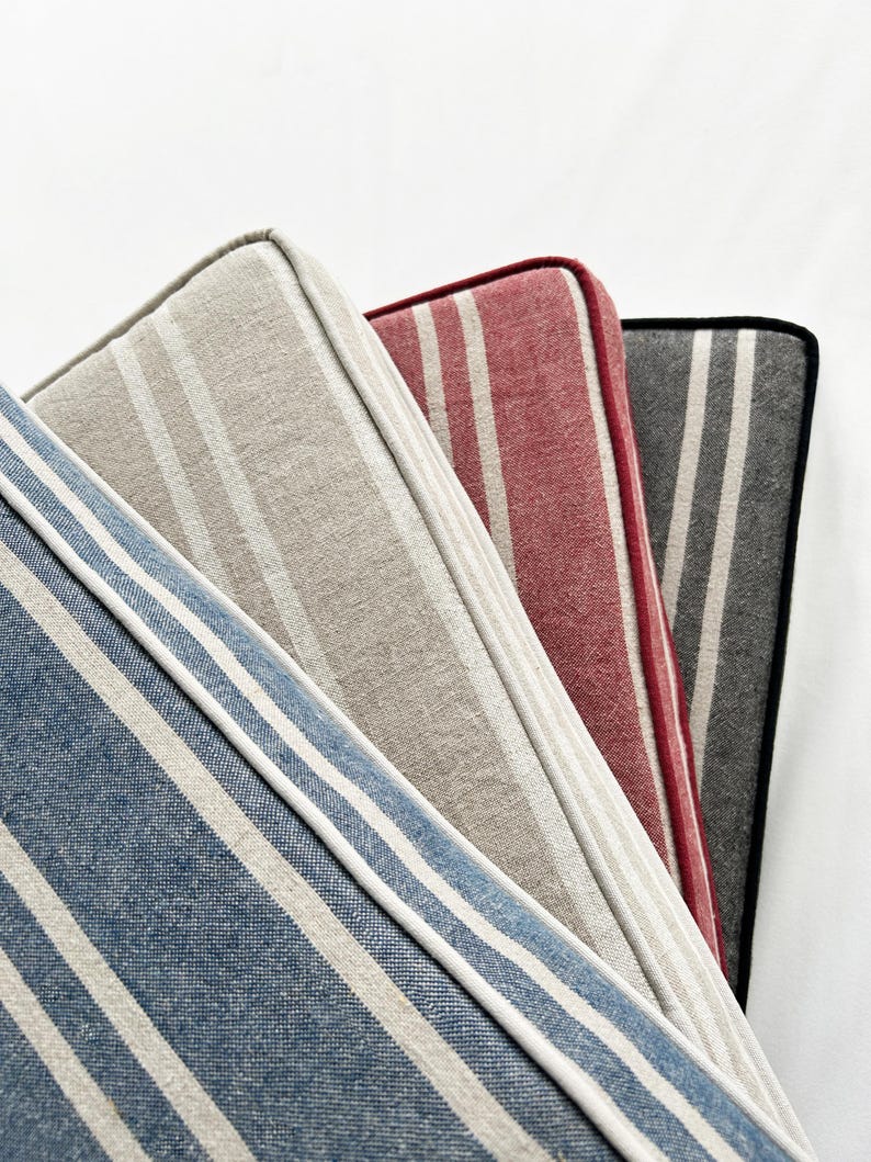 May include: Close-up of four striped cushions in shades of blue, beige, red, and gray. Each cushion has a white border and a different stripe pattern. The cushions are arranged diagonally, showcasing their varying colors and textures.