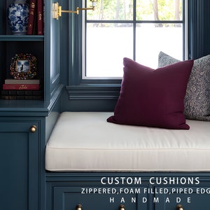 May include: A window seat with a white cushion and two pillows. The seat is built into a dark blue bookcase with a gold knob. The pillows are burgundy and blue with a white pattern. The text "CUSTOM CUSHIONS" is below the seat, and the text "ZIPPERED, FOAM FILLED, PIPED EDGES" is below that. The text "HANDMADE" is below that.