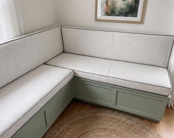 Op de afbeelding: Een L-vormige hoekbank met een saliegroene houten basis met paneeldetails. De bank is voorzien van dikke, lichtbeige stoffen zit- en rugkussens, elk afgewerkt met een dunne, donkergrijze bies langs de randen.