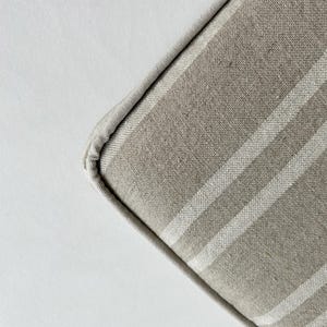 May include: Close-up of a corner of a beige and white striped cushion. The cushion has a light beige fabric with vertical white stripes. The edges are neatly sewn with a matching beige trim. The background is a plain white surface.
