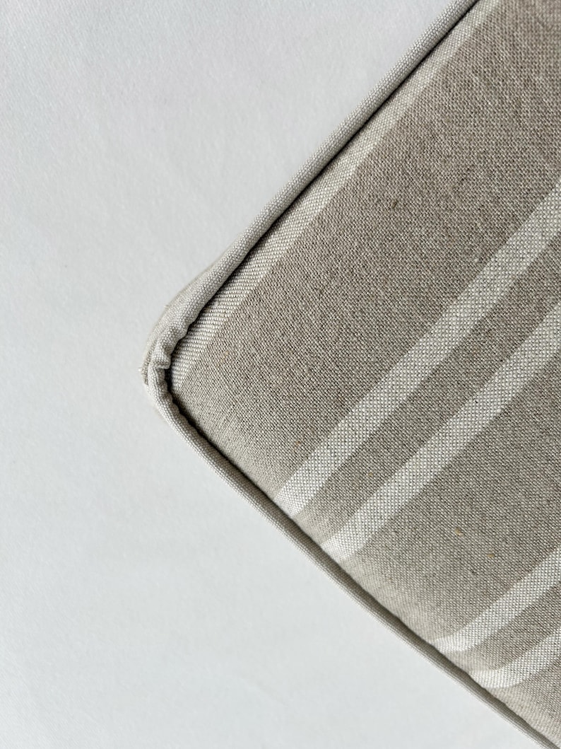 May include: Close-up of a corner of a beige and white striped cushion. The cushion has a light beige fabric with vertical white stripes. The edges are neatly sewn with a matching beige trim. The background is a plain white surface.