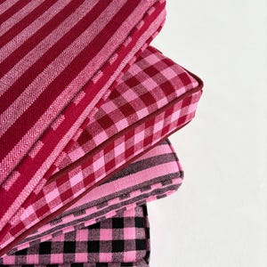 May include: A stack of four square cushions in shades of pink and red. The cushions feature various patterns, including stripes and gingham. Each cushion has a contrasting dark red border.