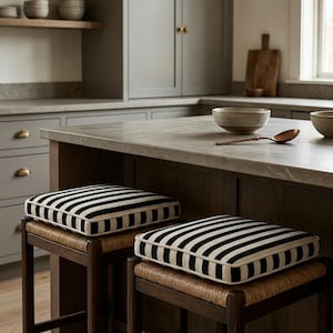 Può includere: Due sgabelli da cucina in legno con sedute intrecciate e cuscini a righe bianche e nere. Gli sgabelli sono posizionati davanti a un'isola cucina con piano in pietra. La cucina ha mobili e mensole grigie.
