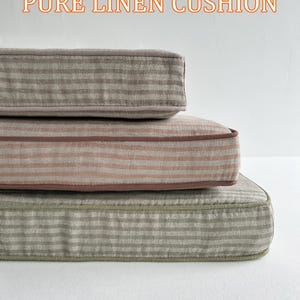 May include: Three stacked linen cushions in shades of gray, pink, and green, each with a striped pattern. The cushions have a piped edge and the text "PURE LINEN CUSHION" in orange.