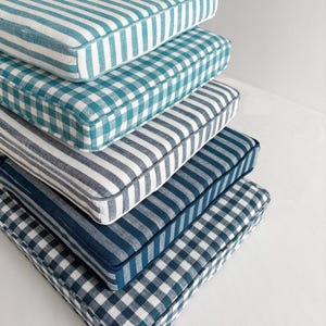 Puede incluir: Una pila de cinco cojines cuadrados con varios patrones azules y blancos. Los patrones incluyen rayas y cuadros de vichy. Cada cojín tiene un borde blanco.