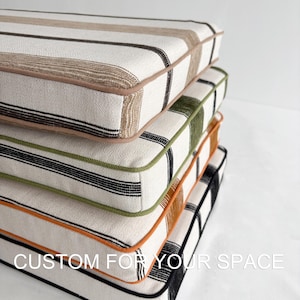 May include: A stack of four square cushions with striped and plaid patterns. The cushions have a white background with stripes in brown, black, and green. Each cushion is trimmed with a contrasting color, and the text "CUSTOM FOR YOUR SPACE" is visible.