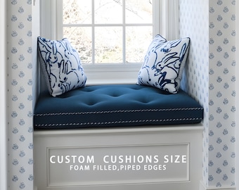Puede incluir: Un cojín de asiento de ventana azul con dos almohadas blancas con un patrón de conejo azul. El cojín está relleno de espuma y tiene bordes con ribete. El texto "CUSTOM CUSHIONS SIZE" y "FOAM FILLED, PIPED EDGES" está impreso debajo del cojín.