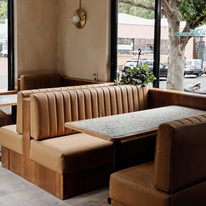 May include: A restaurant booth with tan leather seating and wooden paneling. The booth has a high back with vertical stitching. Tables with speckled countertops are in front of the seating. A window offers a view of the street.