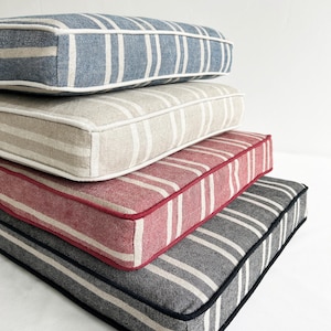 May include: A stack of four rectangular cushions with striped patterns. The top cushion is blue with white stripes and a white border. The next cushion is beige with white stripes and a beige border. The third cushion is red with white stripes and a red border. The bottom cushion is gray with white stripes and a black border.