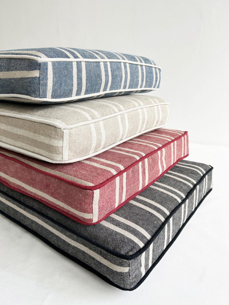 May include: A stack of four rectangular cushions with striped patterns. The top cushion is blue with white stripes and a white border. The next cushion is beige with white stripes and a beige border. The third cushion is red with white stripes and a red border. The bottom cushion is gray with white stripes and a black border.