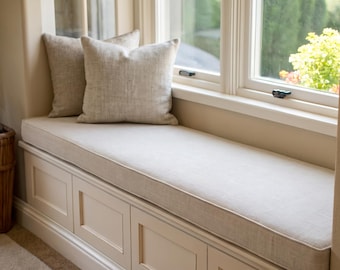 Op de afbeelding: Een vensterbank met een lang, rechthoekig beige kussen met biezen, bekroond met twee vierkante beige sierkussens. De zitting is ingebouwd in een witte houten basis met paneelwerk en bevindt zich direct onder een raam.