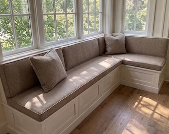 Può includere: Una panca angolare integrata a forma di L con base in legno bianco a pannelli incassati e spessi cuscini di seduta e schienali imbottiti color marrone chiaro. Due cuscini quadrati abbinati sono posizionati sulla seduta della panca.