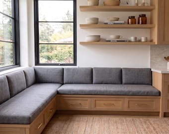 May include: An L-shaped built-in bench with light wood cabinetry and three storage drawers featuring thin gold handles. The bench is topped with thick, dark gray fabric cushions and backrests, positioned against a white wall near a window.