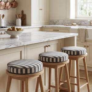 May include: Three wooden bar stools with round seats. The seats have a gray and white striped pattern. The stools are in a kitchen with a marble countertop and cream-colored cabinets. Copper pots hang above the stove.