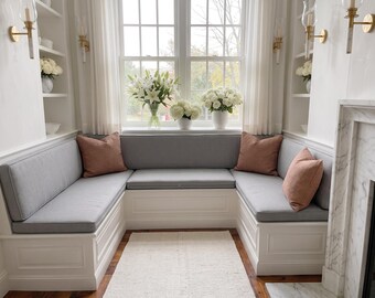 May include: A white built-in U-shaped bench with gray seat cushions and back cushions. Three dusty rose square throw pillows are placed on the bench. The bench is positioned in front of a window with white curtains and white floral arrangements.