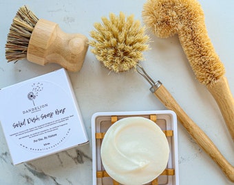 May include: A solid dish soap bar in a white and bamboo soap dish, with two wooden handled dish brushes and a box of soap. The box reads "Dandelion Naturals Solid Dish Soap Bar". The soap is a light cream color.