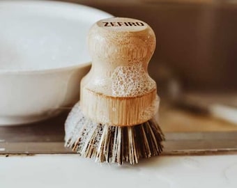May include: A wooden dish brush with the word "ZEFIRO" stamped on the top. The brush has a round handle and bristles. The brush is covered in soap suds. The background includes a white bowl and a stainless steel sink.