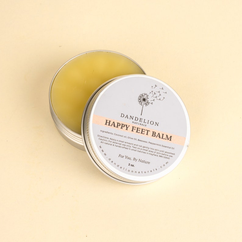 Op de afbeelding: Een ronde, zilveren blik van Happy Feet Balm van Dandelion Naturals, met het deksel iets open. De balsem is lichtgeel van kleur. Het etiket bevat de productnaam, ingredi&euml;nten, instructies en website. De blik bevat 57 gram balsem.