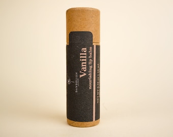 May include: A cylindrical lip balm tube made of cardboard. The tube has a dark gray label with the word "Vanilla" in large white letters. The label also says "nourishing lip balm" and "NET WT 0.33 oz | 10 mL". The brand name "Dandelion" is also printed on the label.