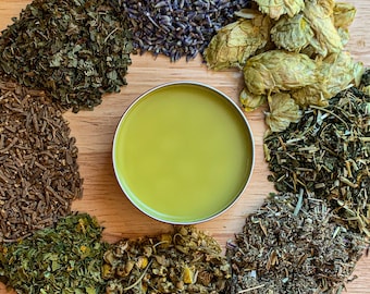 May include: A circular arrangement of dried herbs and flowers surrounds a small, round tin filled with a light green balm. The herbs include lavender, hops, and chamomile, displayed on a wooden surface.