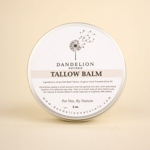 May include: A round, silver-colored tin of Dandelion Naturals Tallow Balm. The label features the text "TALLOW BALM" and a dandelion illustration. The tin contains 2 oz of balm and is made with natural ingredients.