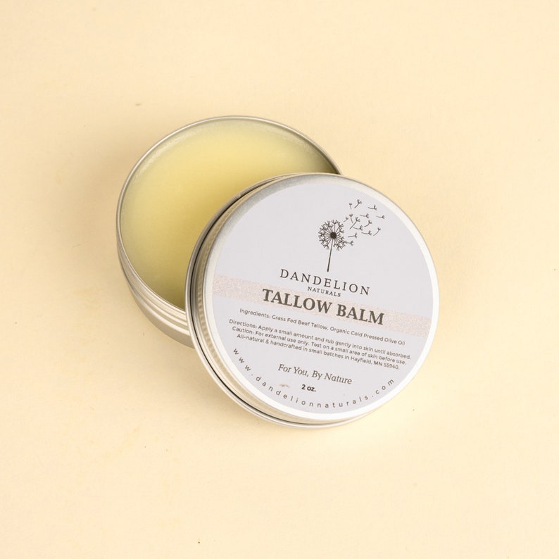 May include: An open, round, silver tin of Dandelion Naturals Tallow Balm. The balm is a light yellow color. The lid is next to the tin, revealing the product and the label. The label has the text "TALLOW BALM" and "2 oz."