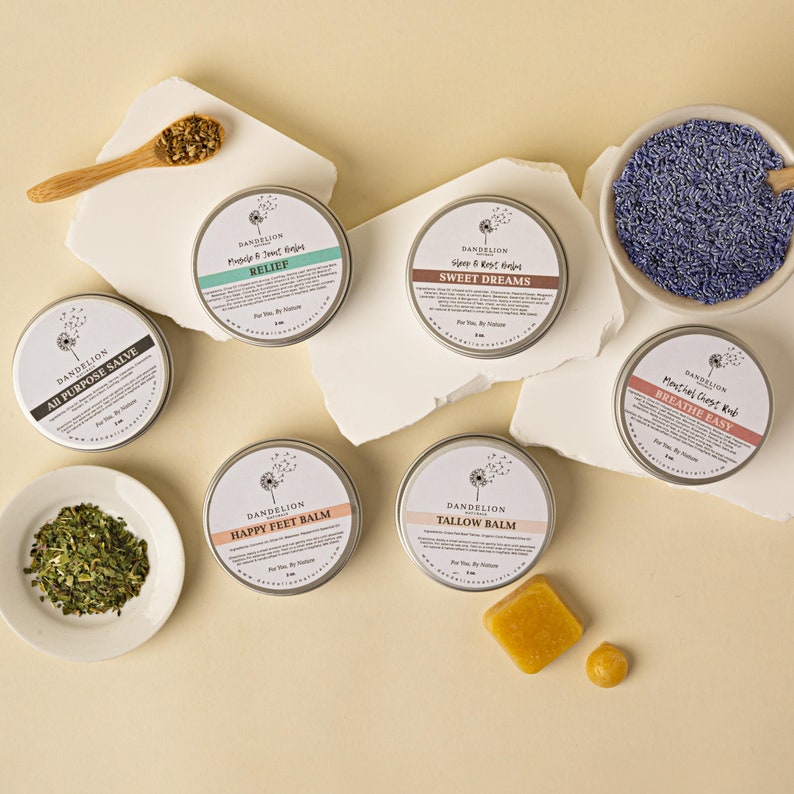 May include: An assortment of Dandelion brand balms in small, round metal tins. The tins are labeled with product names like "Relief", "Sweet Dreams", and "Happy Feet Balm". Accompanying ingredients are displayed in small bowls and a wooden spoon.