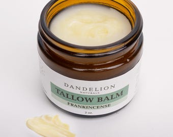 May include: A small, amber-colored glass jar filled with a creamy, pale yellow balm. The jar has a white label with green text that reads "Dandelion Naturals Tallow Balm Frankincense 2 oz." A small smear of balm sits beside the jar.