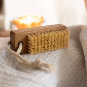 May include: A rectangular wooden nail brush featuring light tan natural bristles and a smooth brown wooden handle. A thick, cream-colored braided cotton rope loop is threaded through a circular hole on one end of the wooden block handle.