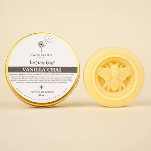May include: A round, light yellow lotion bar with a bee design next to a gold-rimmed tin. The tin lid displays the text "DANDELION NATURALS Lotion Bar VANILLA CHAI" and product information. The lotion bar weighs 1.25 oz.