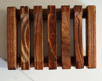 May include: A wooden soap dish with a slatted design. The dish is made of stained wood, featuring vertical slats held together by horizontal supports. The wood grain is visible, and the finish is a warm brown color.