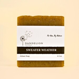 May include: A rectangular bar of artisan soap with a white label featuring a dandelion illustration and the words "Dandelion Naturals" and "Sweater Weather." The soap is a mottled green color and weighs 4-5 oz.