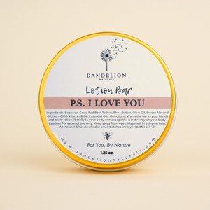 May include: A round lotion bar tin with a gold rim and a white label. The label features the text "Dandelion Naturals Lotion Bar P.S. I Love You." The tin also includes the text "For You, By Nature" and the weight, 1.25 oz. The product is all-natural.