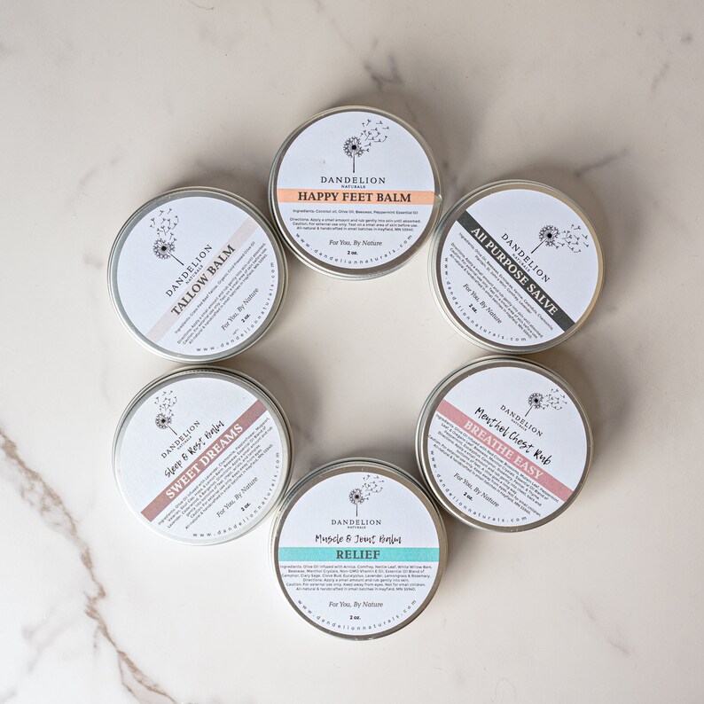 Op de afbeelding: Cirkelvormige metalen blikken met Dandelion Naturals-balsems, gerangschikt in een cirkel. De etiketten tonen productnamen zoals "Happy Feet Balm" en "Sweet Dreams". De blikken zijn zilverkleurig met witte en pastelkleurige etiketten.