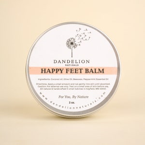 Op de afbeelding: Een ronde, zilverkleurige blik van Happy Feet Balm van Dandelion Naturals. Het etiket toont een paardenbloemillustratie en de tekst "Happy Feet Balm". De balsem bevat kokosolie, olijfolie, bijenwas en essenti&euml;le pepermuntolie. De blik bevat 57 g product.