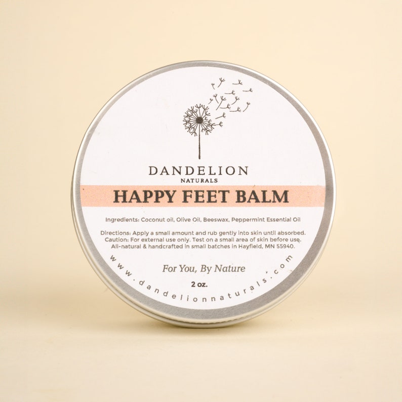 Op de afbeelding: Een ronde, zilverkleurige blik van Happy Feet Balm van Dandelion Naturals. Het etiket toont een paardenbloemillustratie en de tekst "Happy Feet Balm". De balsem bevat kokosolie, olijfolie, bijenwas en essenti&euml;le pepermuntolie. De blik bevat 57 g product.