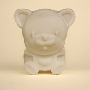 May include: A cream-colored, bear-shaped soap figurine. The bear has rounded features, including ears, a nose, and a seated posture. The soap is a solid, opaque color and appears to be unscented. The background is a light, solid color.