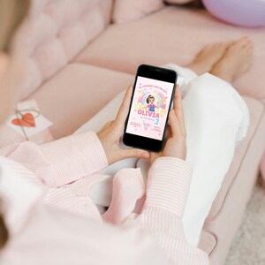 May include: A smartphone displays a birthday invitation with a rainbow, a girl illustration, and the name "Olivia." The invitation includes the number "3" and other event details. The phone is held by a person wearing a pink striped shirt.