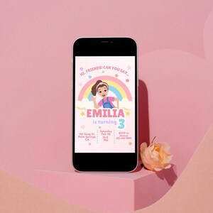 Pastel-themed birthday invitation featuring a cartoon girl inspired by Ms. Rachel, waving with a cheerful expression. The design includes a rainbow, clouds, and colorful stars on a light pink background. The text reads, 'Hi, friends! Can you say…' followed by 'Emilia is turning 3' in bold pink and purple letters. Below, event details such as date, time, location, and RSVP contact are displayed in a playful font. Simply edit, save, and print or share digitally!
