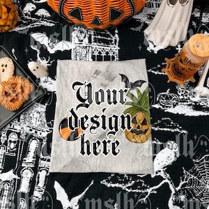 Puede incluir: Una camiseta gris claro con el texto "Your design here" en fuente gótica, junto con un diseño de calabaza y piña. La camiseta se muestra sobre una tela de Halloween en blanco y negro, rodeada de decoraciones de Halloween.