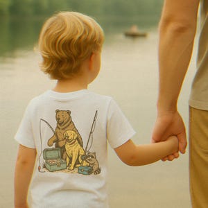 Peut inclure: Un t-shirt blanc avec un dessin d'ours, de chien et de raton laveur p&ecirc;chant. L'enfant tient la main d'un adulte. L'arri&egrave;re-plan est un lac avec un bateau.