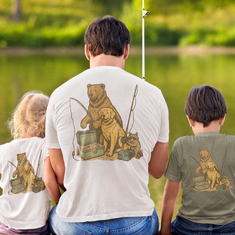 Peut inclure: T-shirts blancs et vert olive avec un motif sur le th&egrave;me de la p&ecirc;che. Le motif repr&eacute;sente un ours, un chien et un raton laveur avec du mat&eacute;riel de p&ecirc;che. L'image montre une famille assise au bord d'un lac, portant des chemises assorties.