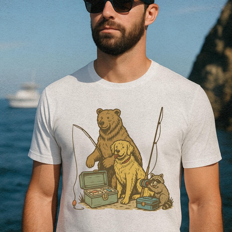 Peut inclure: T-shirt gris clair avec une illustration de dessin anim&eacute; d'un ours, d'un golden retriever et d'un raton laveur p&ecirc;chant. L'ours et le chien sont assis, le raton laveur tenant un moulinet. Une bo&icirc;te &agrave; p&ecirc;che verte est ouverte, et des cannes &agrave; p&ecirc;che sont pr&eacute;sentes.