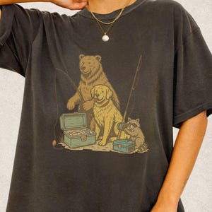 Peut inclure: T-shirt gris fonc&eacute; avec un motif de p&ecirc;che. L'illustration repr&eacute;sente un ours, un chien et un raton laveur, tous avec des cannes &agrave; p&ecirc;che et des bo&icirc;tes &agrave; p&ecirc;che. Le dessin utilise des couleurs marron, vert et bleu. Le t-shirt est en mati&egrave;re douce.