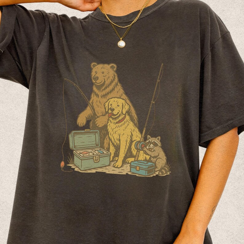 Peut inclure: T-shirt gris fonc&eacute; avec un motif de p&ecirc;che. L'illustration repr&eacute;sente un ours, un chien et un raton laveur, tous avec des cannes &agrave; p&ecirc;che et des bo&icirc;tes &agrave; p&ecirc;che. Le dessin utilise des couleurs marron, vert et bleu. Le t-shirt est en mati&egrave;re douce.