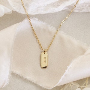 May include: A gold chain necklace with a rectangular pendant that says "heir" in a script font.