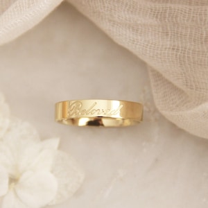 Puede incluir: Un anillo dorado con la palabra "Beloved" grabada en la banda. El anillo se muestra sobre un fondo suave y neutro con flores blancas y tela drapeada.