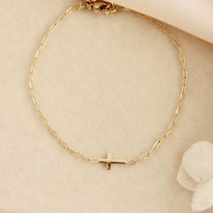 May include: A delicate gold-colored bracelet featuring a small cross pendant. The bracelet has a chain link design and a clasp closure. The cross is centered on the bracelet, adding a touch of elegance. The bracelet is displayed on a neutral background.