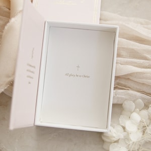 May include: A white box with a pink interior. The box is open and the inside is lined with white paper. The paper has a gold cross and the text "All glory be to Christ" printed on it.